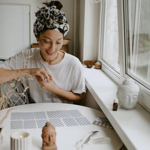 May include: A person sits at a table with a white tablecloth and reads tarot cards. The cards are laid out in a spread on the table. The person is wearing a white shirt and a patterned headscarf. There is a small white Buddha statue on the windowsill behind the person.