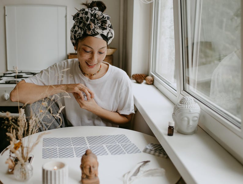 May include: A person sits at a table with a white tablecloth and reads tarot cards. The cards are laid out in a spread on the table. The person is wearing a white shirt and a patterned headscarf. There is a small white Buddha statue on the windowsill behind the person.