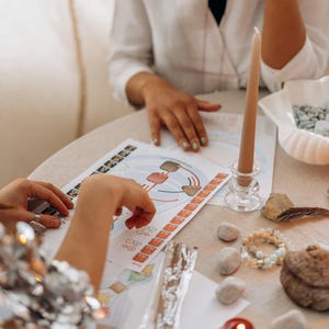 May include: Two people are sitting at a table with a white tablecloth. They are working on a colorful chart with a diagram and numbers. There are candles, stones, and other objects on the table.