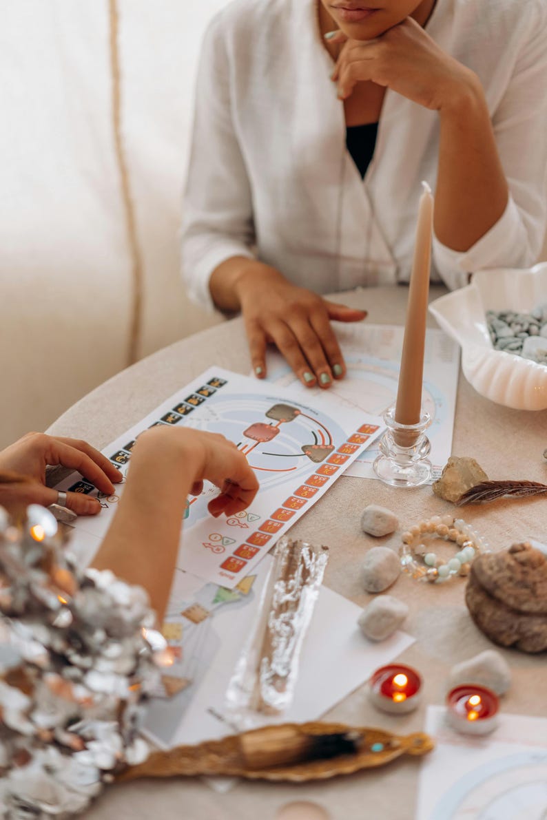 May include: Two people are sitting at a table with a white tablecloth. They are working on a colorful chart with a diagram and numbers. There are candles, stones, and other objects on the table.
