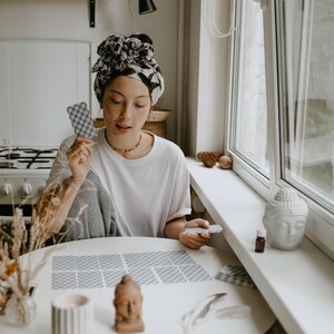 May include: A person seated at a round white table, arranging tarot cards. The person wears a white t-shirt and a patterned headscarf. A small Buddha statue and a lit candle are on the table. A window and a stove are in the background.