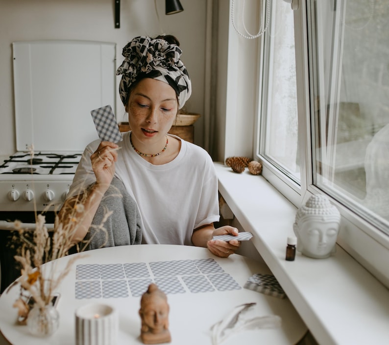 May include: A person seated at a round white table, arranging tarot cards. The person wears a white t-shirt and a patterned headscarf. A small Buddha statue and a lit candle are on the table. A window and a stove are in the background.