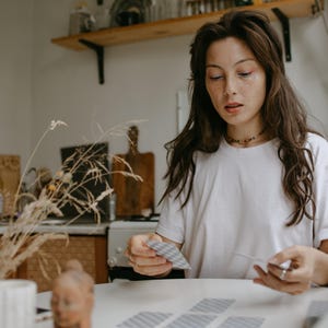 May include: A woman with long brown hair sits at a table, holding a deck of tarot cards. She is wearing a white t-shirt and has freckles on her face. The table is white and there are several tarot cards laid out on it.