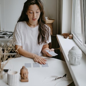 May include: A person is seated at a white table, arranging tarot cards. A small Buddha statue and a lit candle are on the table. The person is wearing a white t-shirt and a beaded necklace. A white Buddha head statue sits on a windowsill.