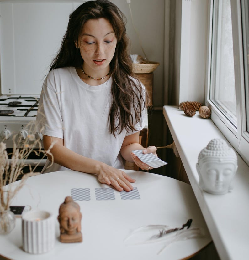 May include: A person is seated at a white table, arranging tarot cards. A small Buddha statue and a lit candle are on the table. The person is wearing a white t-shirt and a beaded necklace. A white Buddha head statue sits on a windowsill.