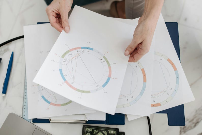 May include: A person is holding a white sheet of paper with a colorful circle chart on it. The chart has a blue, green, pink, and orange ring around the outside. There are lines and numbers inside the circle. The person is holding the paper over other similar sheets of paper with circle charts.