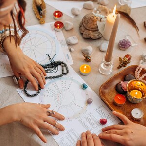 May include: Two people are looking at a white astrological chart with a circle in the center and a grid of numbers around it. The chart is on a white tablecloth with candles, crystals, and other objects around it.