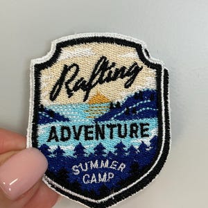 Puede incluir: Parche bordado con forma de escudo. El parche presenta las palabras "Rafting Adventure Summer Camp" con un diseño de paisaje de río, árboles y puesta de sol. El parche está bordeado con un borde cosido en blanco y negro.