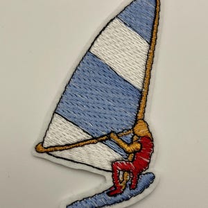 Könnte beinhalten: Ein gestickter Aufnäher mit einer Person beim Windsurfen. Die Person trägt einen roten Neoprenanzug und hält ein goldenes Segel mit blauen und weißen Streifen. Der Aufnäher ist auf weißem Hintergrund.