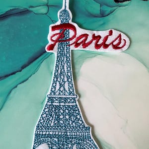 Könnte beinhalten: Weißer gestickter Aufnäher mit einer blauen Umrandung des Eiffelturms und dem Wort "Paris" in roter Kursivschrift.