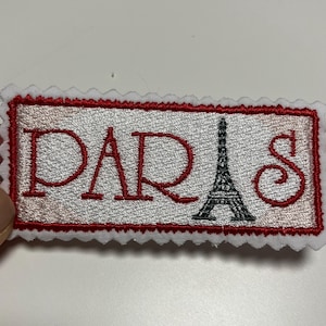 Könnte beinhalten: Gestickter Aufnäher mit dem Wort "PARIS" in Rot und einem schwarzen Eiffelturm-Bild. Der Aufnäher hat einen weißen Hintergrund, einen roten Rand und einen gewellten Rand.