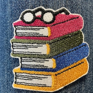 Könnte beinhalten: Gestickter Aufnäher mit einem Stapel Bücher in Rosa, Grün, Blau und Gelb, gekrönt von einer Brille mit schwarzem Rahmen. Der Aufnäher ist mit weißem Faden gestickt und hat einen weißen Rand. Der Aufnäher befindet sich auf einem Jeans-Hintergrund.