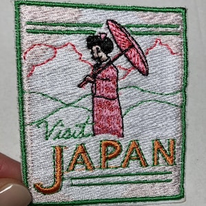 May include: Embroidered patch featuring a geisha in a pink kimono holding a red parasol. The patch reads "Visit JAPAN" in orange letters, set against a white background with green and red accents. The patch is square-shaped.