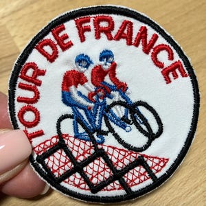 Könnte beinhalten: Runder, gestickter Aufnäher mit den roten Worten "TOUR DE FRANCE" auf weißem Hintergrund. Der Aufnäher zeigt zwei Radfahrer in Rot und Blau auf einem Fahrrad. Ein schwarzer Rand umgibt den Aufnäher.