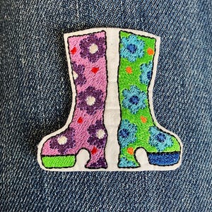 Könnte beinhalten: Gestickter Aufnäher mit einem Paar bunter Stiefel. Ein Stiefel ist rosa mit lila Blumen, der andere ist grün mit blauen Blumen. Beide Stiefel haben einen weißen Rand und sind vor einem Jeanshintergrund platziert. Die Stiefel haben einen Plateauabsatz.