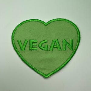 Könnte beinhalten: Ein grüner, herzförmiger, gestickter Aufnäher mit dem Wort "VEGAN" in grüner Farbe. Der Aufnäher hat einen gestickten Rand und ist vor einem weißen Hintergrund platziert. Ideal für Kleidung oder Accessoires.