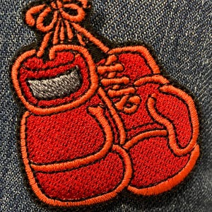 Könnte beinhalten: Rote gestickte Boxhandschuhe, die mit einer schwarzen Schnur zusammengebunden sind. Die Handschuhe befinden sich auf einem blauen Jeansstoff.