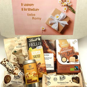 Può includere: Una scatola regalo piena di vari snack e dolcetti vegani, tra cui barrette di cioccolato, marzapane, palline energetiche e tè. La scatola è decorata con un motivo floreale rosa e bianco e le parole "Happy Birthday liebe Romy".