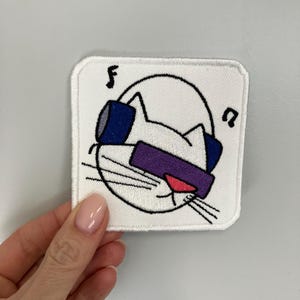 Könnte beinhalten: Weißer gestickter Patch mit einer Comic-Katze, die Kopfhörer und Sonnenbrille trägt. Die Katze hört Musik und hat eine rosa Nase. Der Patch hat einen weißen Hintergrund mit schwarzen Nähten an den Rändern.