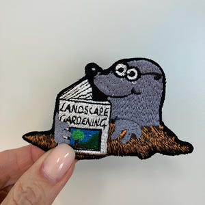 Könnte beinhalten: Gestickter Aufnäher mit einem Cartoon-Maulwurf, der eine Brille trägt und ein Buch mit dem Titel "Landscape Gardening" liest. Der Maulwurf ist grau mit einem braunen Erdfleck. Das Buch hat ein grünes und blaues Bild.