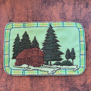 Könnte beinhalten: Gestickter Aufnäher mit einer Hüttenlandschaft mit immergrünen Bäumen. Die Hütte ist braun und die Bäume sind dunkelgrün. Der Aufnäher hat einen hellgrünen Hintergrund mit einem blau-gelben Rand. Der Aufnäher ist rechteckig.