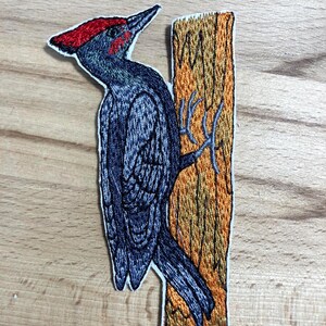 Könnte beinhalten: Gestickter Aufnäher mit einem Specht mit rotem Kamm, der auf einem Baumstamm sitzt. Der Vogel ist überwiegend dunkelblau mit detaillierten Federn. Der Baumstamm ist braun und orange.
