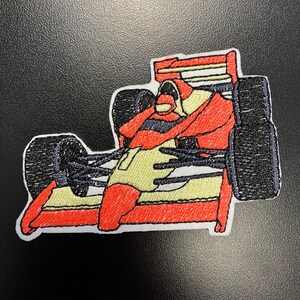 Könnte beinhalten: Gestickter Aufnäher mit einem roten Rennwagen und einem Fahrer in einem gelben Overall. Das Auto hat schwarze Räder und einen weißen Hintergrund.