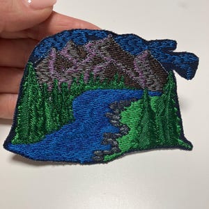 Könnte beinhalten: Gestickter Aufnäher mit einer Berglandschaft mit blauem Fluss, grünen Bäumen und lila Bergen unter einem dunkelblauen Himmel. Der Aufnäher hat einen dunkelblauen Rand und ist für Kleidung oder Accessoires geeignet.