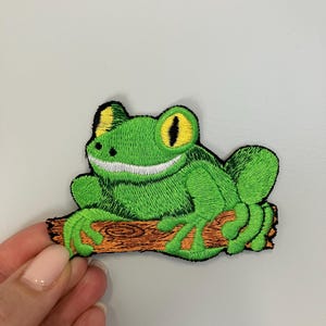 May include: Embroidered patch of a green frog with yellow eyes and a white belly, perched on a brown log. The frog patch has a detailed, textured appearance, with a black outline.