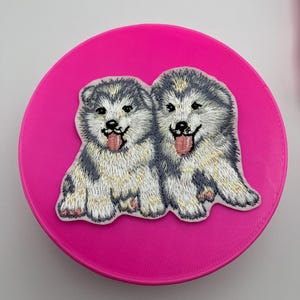 Könnte beinhalten: Gestickter Aufnäher mit zwei weißen und grauen Husky-Welpen mit rosa Nasen und herausgestreckten Zungen. Der Aufnäher befindet sich auf einem rosa Hintergrund.