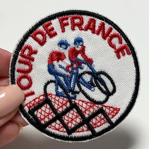 Könnte beinhalten: Gestickter Aufnäher mit dem Schriftzug "TOUR DE FRANCE" in Rot, der ein Design von zwei Radfahrern in Rot und Blau auf Fahrrädern umrahmt. Der Aufnäher hat einen weißen Hintergrund mit einem schwarzen Rand.