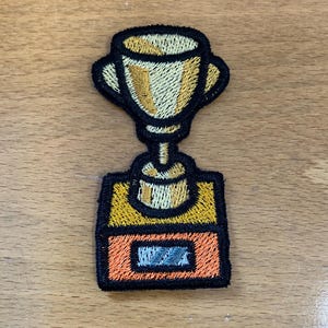 May include: Embroidered patch featuring a gold trophy with a black outline. The trophy sits on a rectangular base with orange and yellow sections, and a small blue rectangle. The patch is designed for application on clothing or accessories.