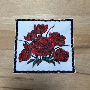 May include: Embroidered patch featuring a bouquet of red roses with green leaves on a white background. The patch has a black braided border. The roses are detailed with various shades of red, creating a realistic appearance. The patch is square-shaped.