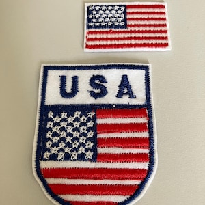 Puede incluir: Dos parches bordados con la bandera estadounidense. Uno es rectangular, el otro en forma de escudo con "USA" en azul. Ambos tienen un borde blanco, rayas rojas y blancas y un campo azul con estrellas blancas. Adecuado para ropa.