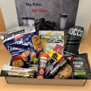 Op de afbeelding: Een geschenkdoos gevuld met fitness- en gezondheidsproducten. Bevat eiwitchips, eiwitrepen, supplementen, een blikje NOCCO-limonade en een eiwitkoekje. De doos heeft de tekst "No Pain. No Gain."