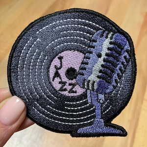 Könnte beinhalten: Gestickter Aufnäher mit einer Vinyl-Schallplatte und einem Mikrofon. Die Schallplatte ist dunkelblau mit weißen Nähten und einem lavendelfarbenen Mittelteil mit dem Wort "JAZZ". Das Mikrofon ist lila und weiß.