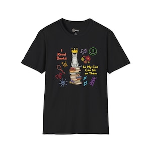 May include: Black t-shirt featuring a cat wearing a crown, perched on books. The text reads "I Read Books So My Cat Can Sit on Them", with colourful doodles surrounding the image.