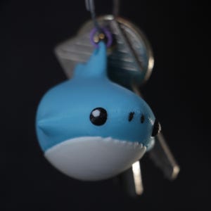 May include: A blue and white shark keyring with black eyes and a small fin. The keyring is attached to a purple ring and a set of keys. The shark has a cartoonish design.