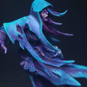 Puede incluir: Figura en miniatura de una criatura fantástica con una capa con capucha. La figura está pintada en tonos azules y morados, con detalles intrincados y una expresión amenazante. El tema general es la fantasía y el arte en miniatura.