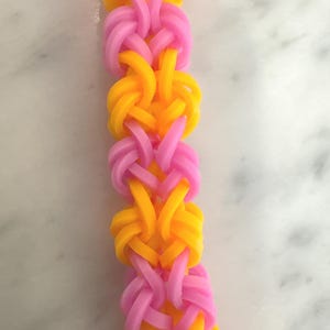 May include: A handmade bracelet featuring a woven design of yellow and pink rubber bands. The bracelet is made with a series of interlocking loops, creating a textured and colorful accessory. The bright colors make it a fun and playful item.