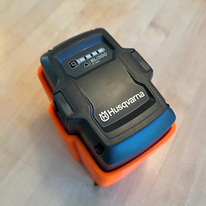 Op de afbeelding: Een zwarte Husqvarna BLJ300 accu met een laadindicator die een volle lading aangeeft. De accu bevindt zich in een oranje kunststof houder.