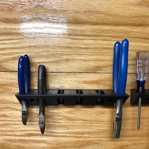 May include: A black plastic tool holder with five slots mounted on a wooden wall. The holder has two pairs of blue pliers and a pair of black pliers. There are also two screwdrivers with black and yellow handles.