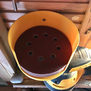 Può includere: Un contenitore di stoccaggio in plastica giallo con un mucchio di dischi abrasivi marroni all'interno. Il contenitore ha un foro nella parte superiore per appenderlo. Una levigatrice elettrica nera e gialla è visibile sullo sfondo.