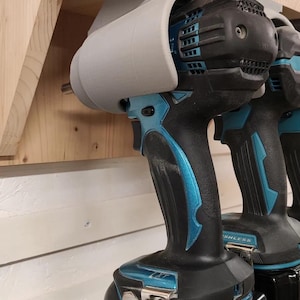 Può includere: Due trapani a batteria blu e neri con supporti grigi montati su uno scaffale di legno. I trapani hanno batterie nere con la scritta "Makita 5.0Ah Li-ion" su di esse.
