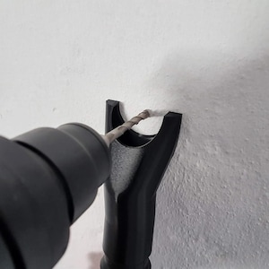 Puede incluir: Una broca negra se inserta en una herramienta negra, perforando una pared blanca. La broca es gris y tiene un diseño en espiral. La herramienta se sujeta contra la pared, creando un agujero.