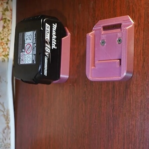 Könnte beinhalten: Eine rosa 3D-gedruckte Wandhalterung für einen Makita 18V 5.0Ah Lithium-Ionen-Akku. Die Halterung hat zwei Schraubenlöcher zur Befestigung an einer Wand.