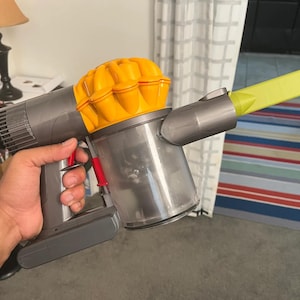 Puede incluir: Una aspiradora inalámbrica de mano con un diseño amarillo y gris. La aspiradora tiene un depósito de polvo transparente y una herramienta de ranura amarilla.