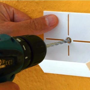 May include: A white plastic template with a crosshair pattern and a hole in the centre. A drill bit is being used to drill a hole through the template. The drill is green and black.