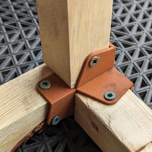 May include: Close-up of a corner joint for a wooden structure. The joint is reinforced with a brown plastic corner brace and secured with screws.