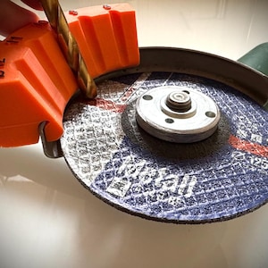 Peut inclure: Guide d'angle en plastique orange pour meuleuse avec un foret inséré. Le guide est marqué pour différents angles. La meuleuse a un disque abrasif bleu et noir avec le texte "Norton" dessus.